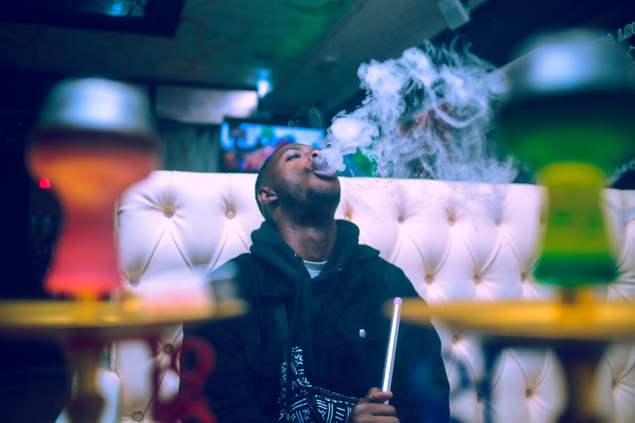 A young man relaxing and smoking shisha at an indoor lounge, surrounded by colorful hookahs.